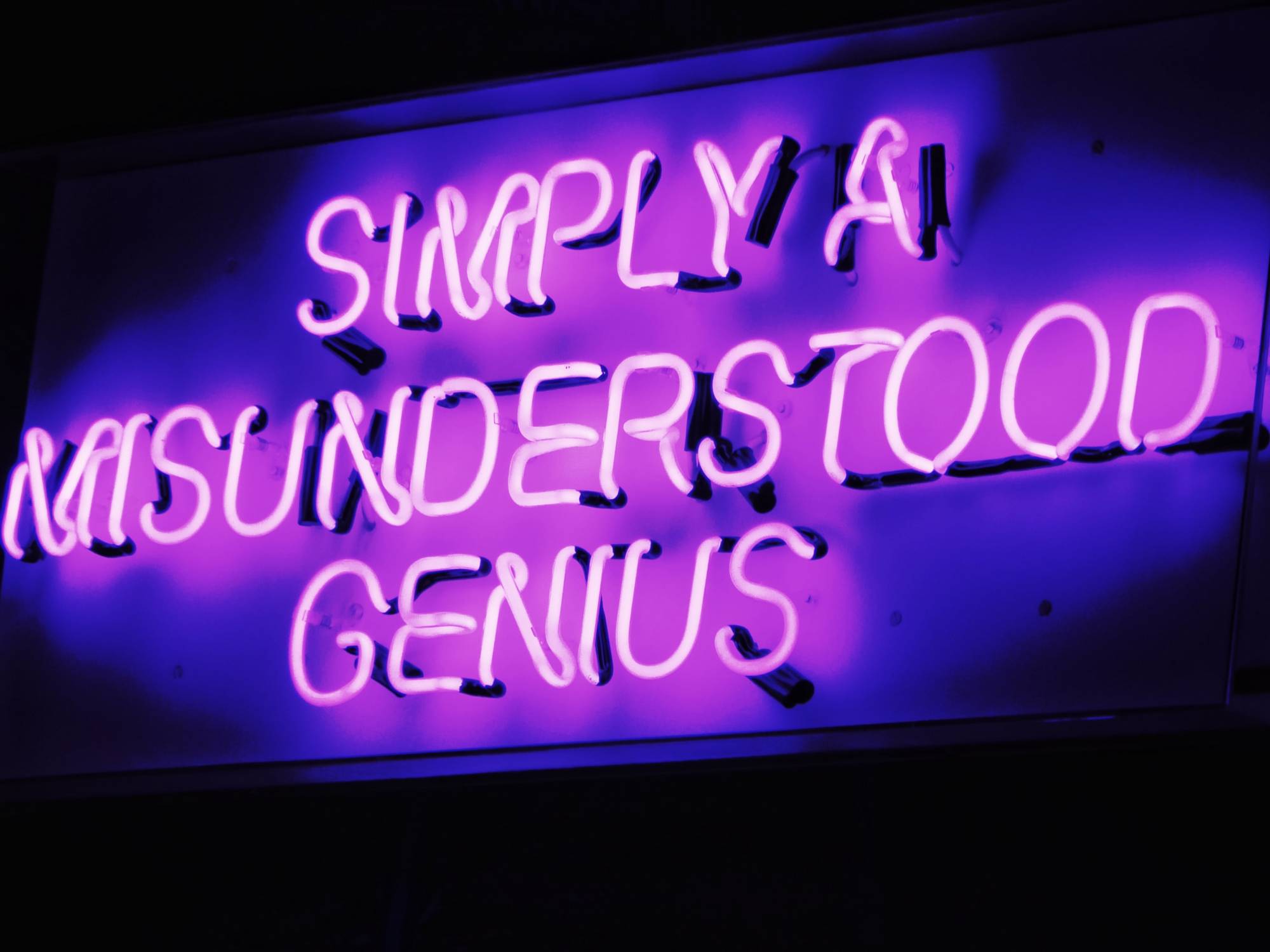 A photo of a purple neon sign displaying text "Simply A Misunderstood Genius"