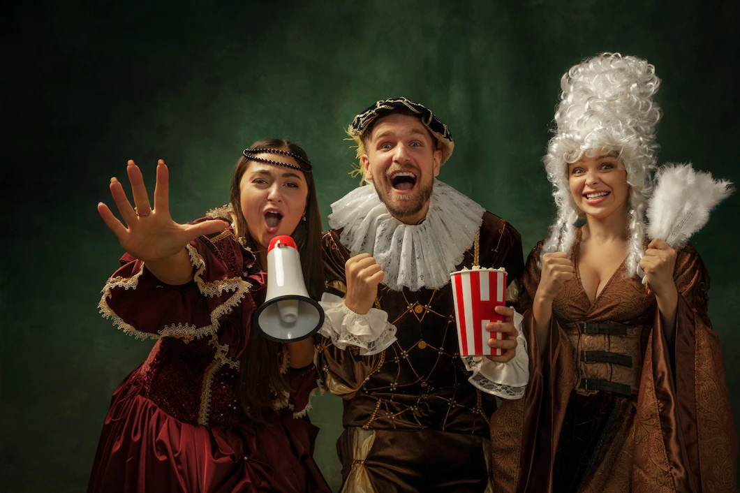 Three people in renaissance wardrobe stand in front of a mottled green background. They are looking towards the camera excitedly, one is holding a megaphone, another a bag of popcorn.