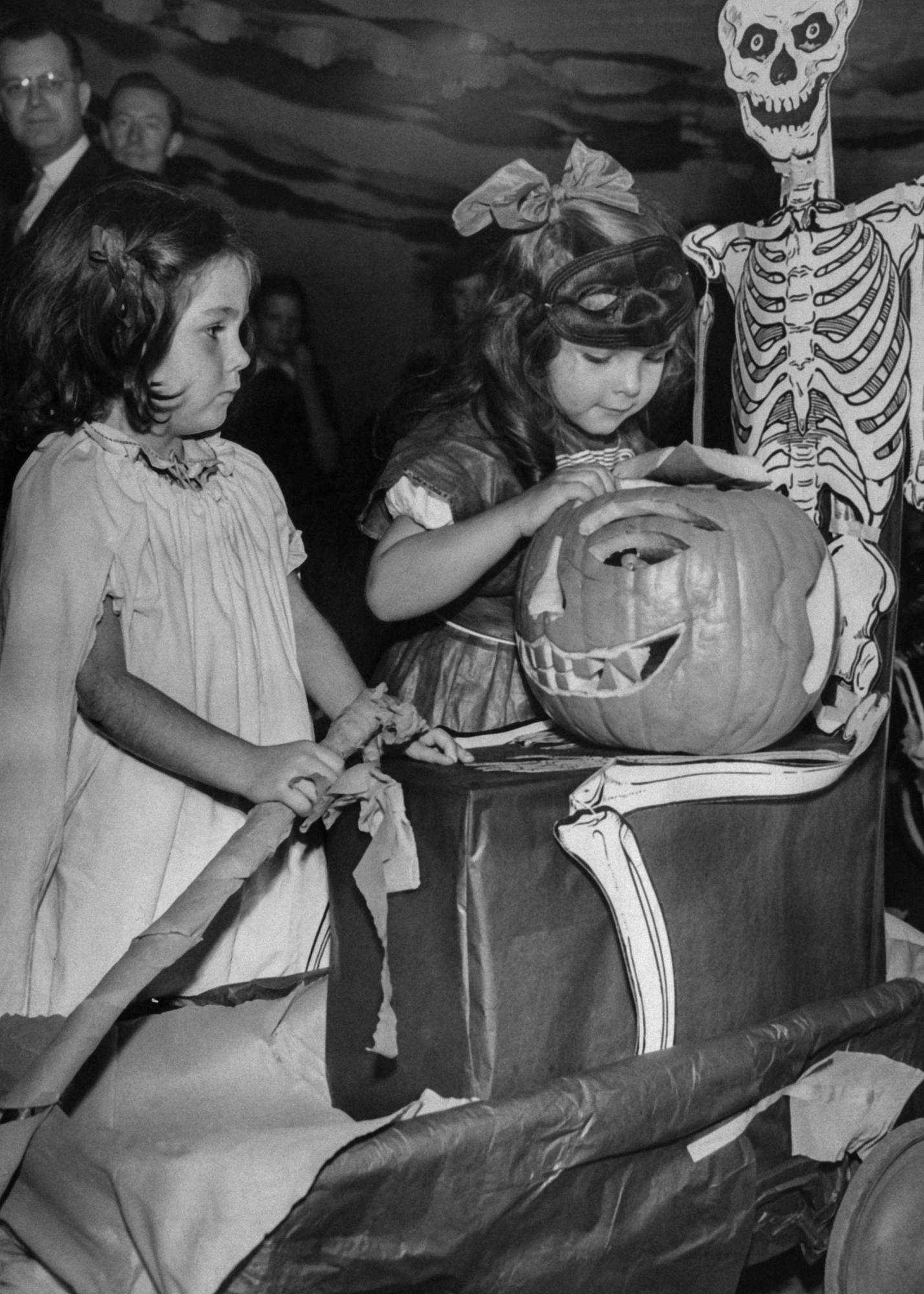 A black and white photo of two girls at an old halloween party with an uncomfortably detailed jack o' lantern. Its creepy, but I don't know why.