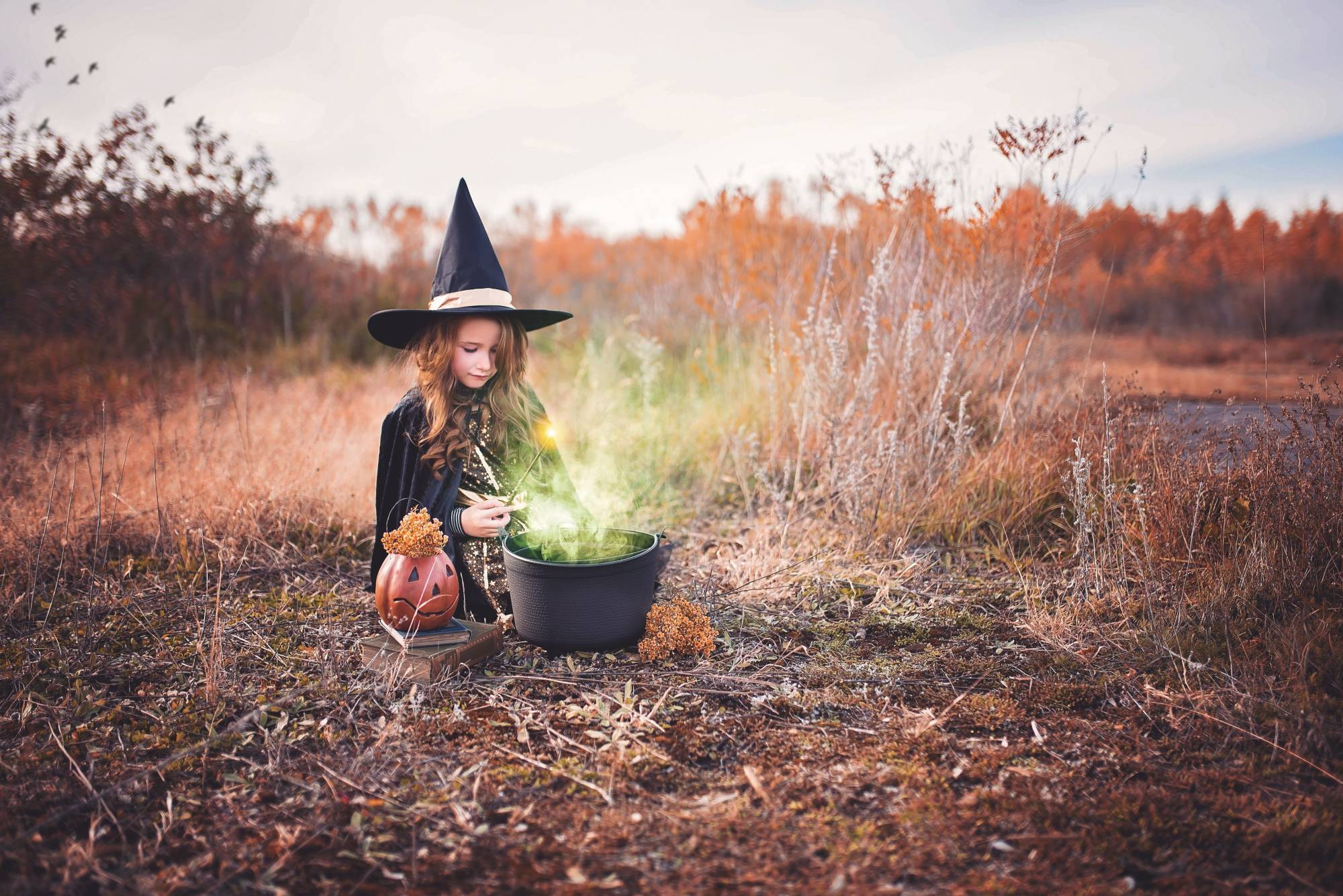 A girl dressed in a witch costume sitting by a black cauldron.