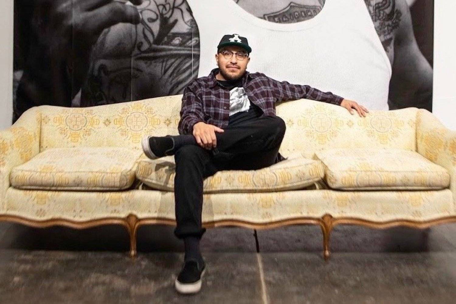 A photo of documentary photographer Juan Fuentes, sitting in the center of a yellow upholstered couch. There is a large-scale floor-to-ceiling black and white image on the wall behind him.