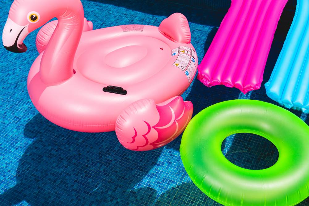 A photo of brightly colored pool floaties in a bright blue pool. Floaties include a pink flamingo, neon green inner tube, hot pink lounge floaty and aqua lounge floaty.