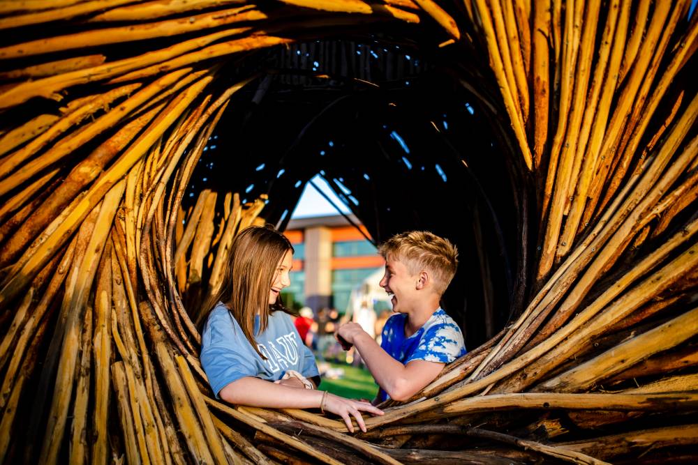 One teenage girl and one boy laugh while standing inside the Anythink Wright Farms wood spirit nest. They are both wearing blue short sleeve shirts.