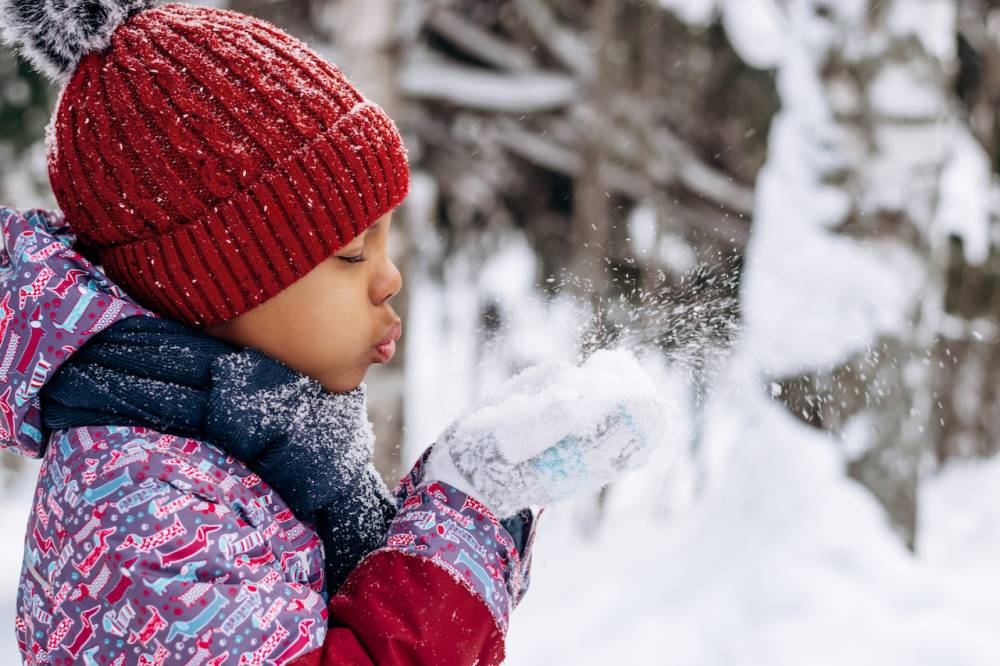 A young girl blows a pile of snow from her hands. She's dressed in winter gear and is outside in the snow.