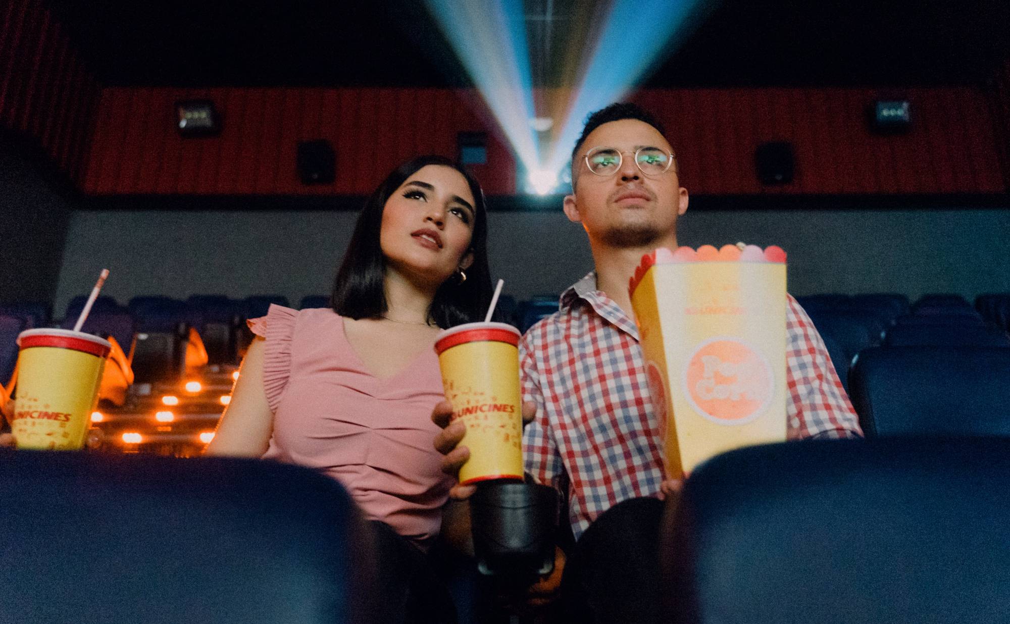 Two people sit watching a movie in a theater while eating popcorn and drinking soda.