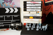 Image of a desk covered in various paraphernalia. On the left, a stack of VHS tapes sit behind miniature game figurines. On the right is a clapboard, a vintage camera, and a pair of tickets.