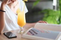 A photo of a woman sitting at a wood table and reading a large book with a orange cocktail and a iPhone in front of her. She is wearing a white t-shirt. The drink is in a tropical cocktail glass with a straw, swirled garnish and citrus fruit slice garnish.The photo shows her neck and torso.