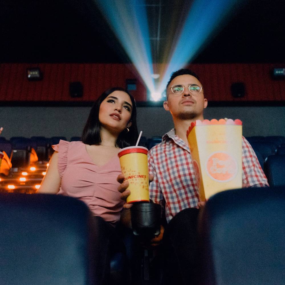 Two people sit watching a movie in a theater while eating popcorn and drinking soda.